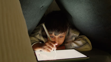 Young-Boy-In-Home-Made-Camp-Made-From-Cushions-Playing-And-Drawing-With-Digital-Tablet-And-Stylus-At-Night